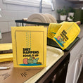 Load image into Gallery viewer, A yellow dishcloth with the text 'SHIT HAPPENS SOAK IT UP' and a cactus illustration stands on a kitchen counter next to other matching dishcloths. Plates and a sink are visible in the background.