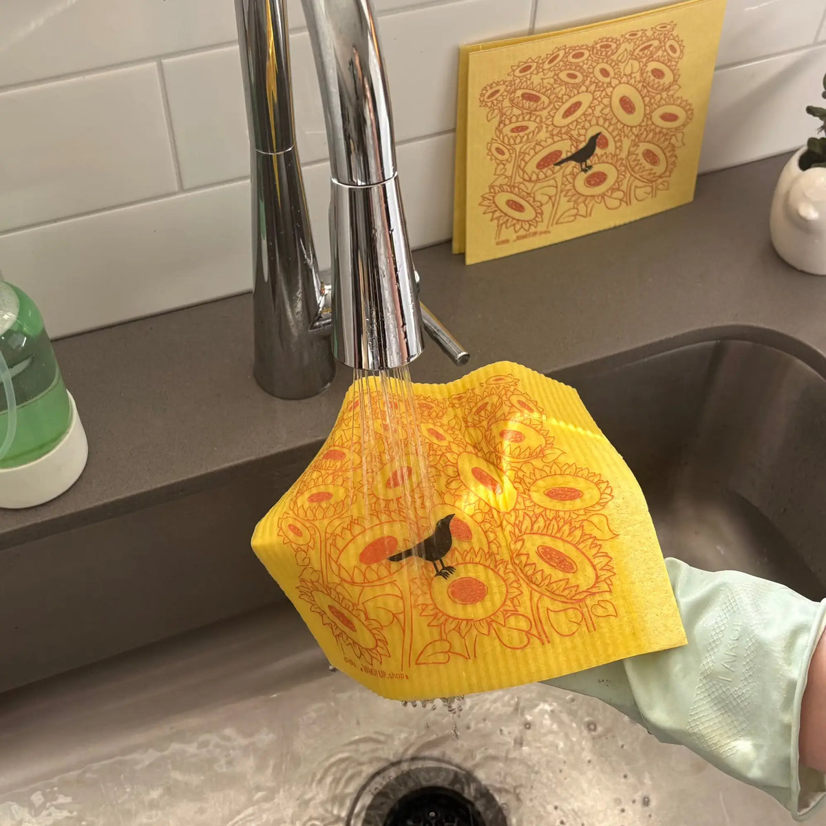 A yellow swedish dishcloth washed under running water in a stainless steel kitchen sink. A hand wearing a light green glove holds the scrubber.
