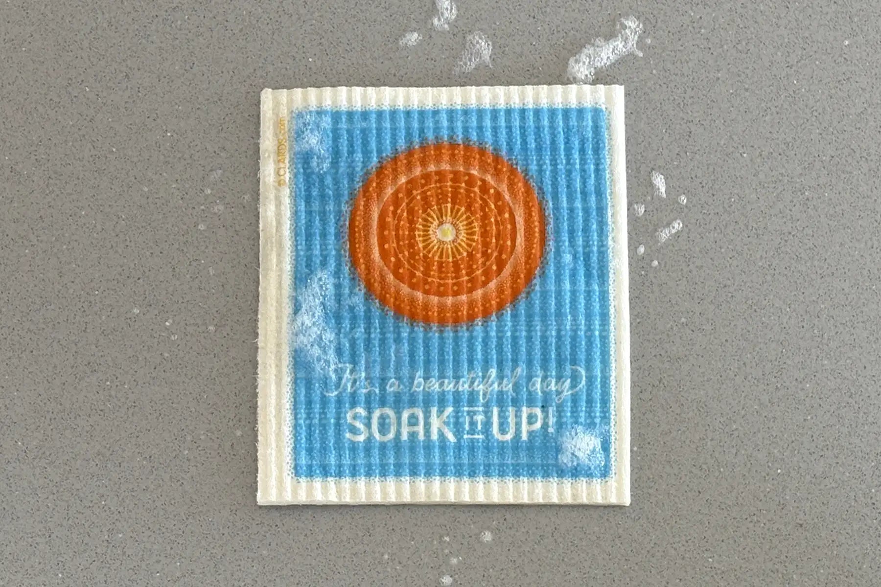 Blue and orange patterned dishcloth with a mandala design and the text ’it’s a beautiful day soak up!’