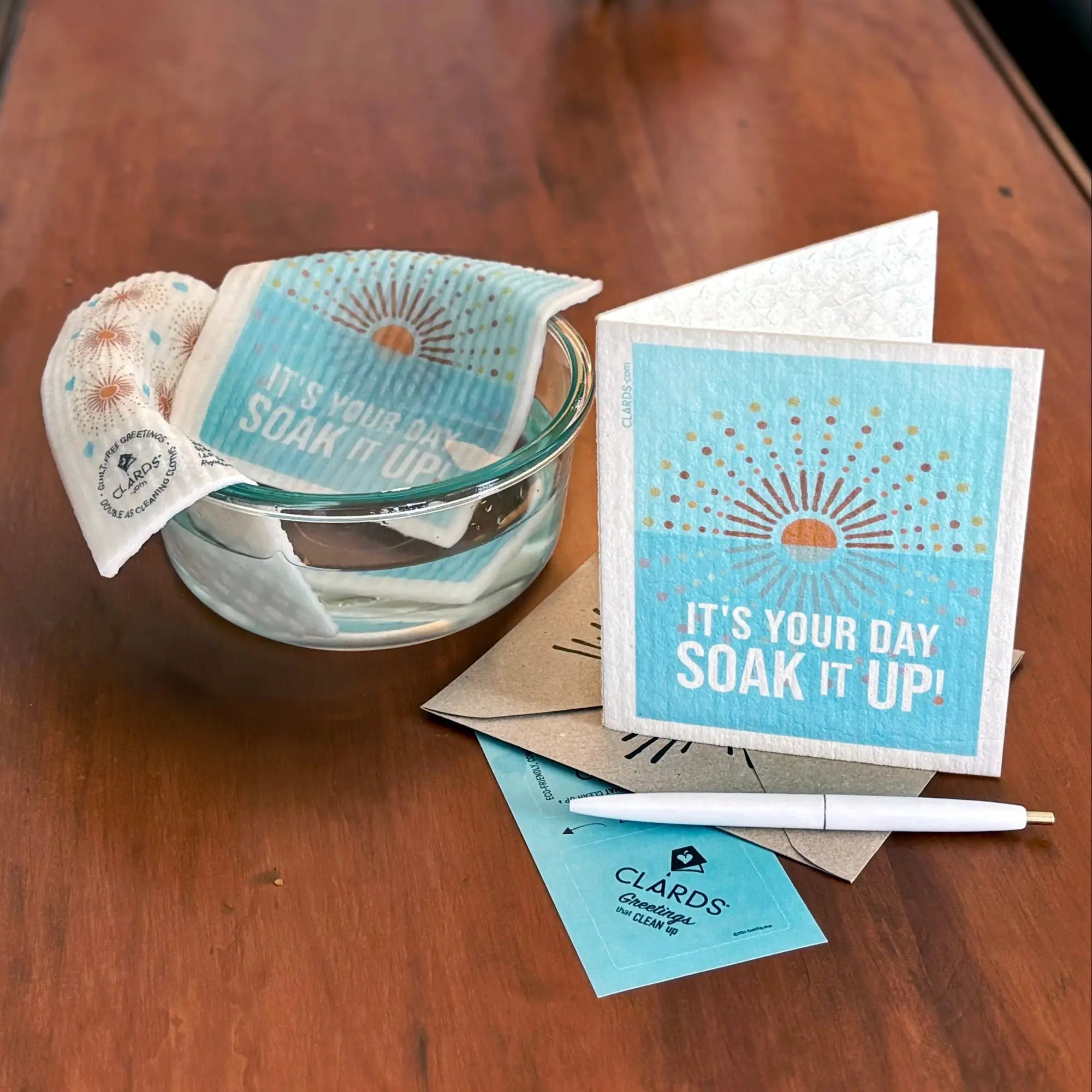 Greeting Clards on wood table showing greeting, pen and envelope and bowl of water with Clards turned into a cleaning cloth. It's Your Day Soak iT Up. soakitup.shop