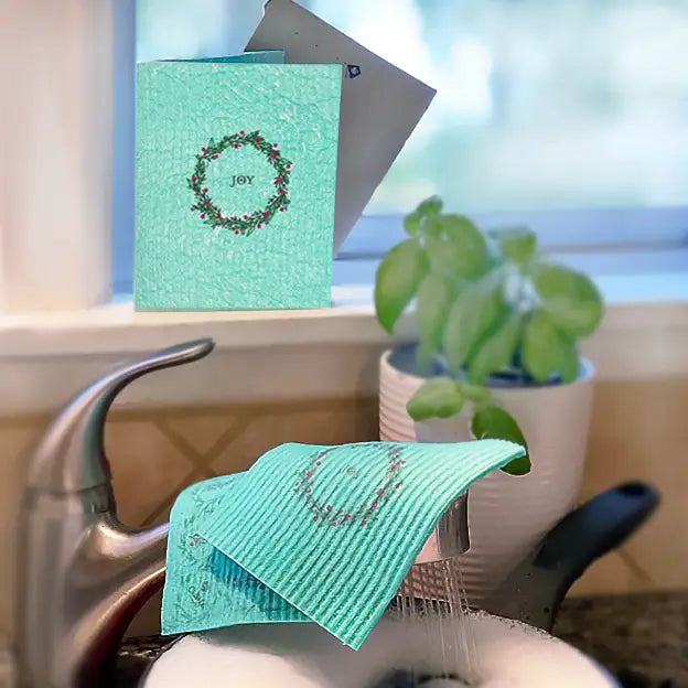 Teal ribbed eco friendly cleaning cloth with subtle joy holiday wreath design resting on sink edge