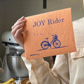 Load image into Gallery viewer, Joy rider bicycle bright orange Swedish dishcloth held by a hand in kitchen setting. Soakitup.shop

