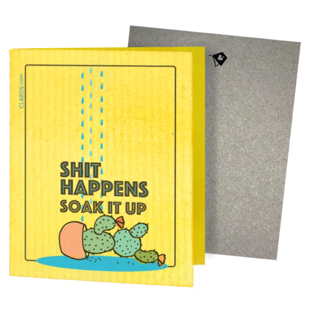 A yellow rectangular card or box with a humorous printed design reading 'SHIT HAPPENS SOAK IT UP' above a cartoon cactus in a puddle. Greeting turns into cleaning cloths