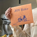 Load image into Gallery viewer, Joy rider bicycle bright orange Swedish dishcloth held by a hand in kitchen setting. Soakitup.shop
