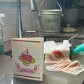 Load image into Gallery viewer, Kitchen counter with a compost bin, cleaning supplies, and a card with a cake illustration. greeting card turns into a cleaning cloth
