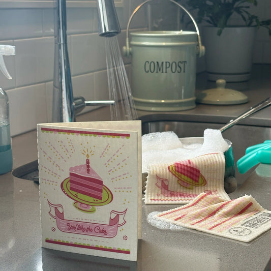 Kitchen counter with a compost bin, cleaning supplies, and a card with a cake illustration. greeting card turns into a cleaning cloth